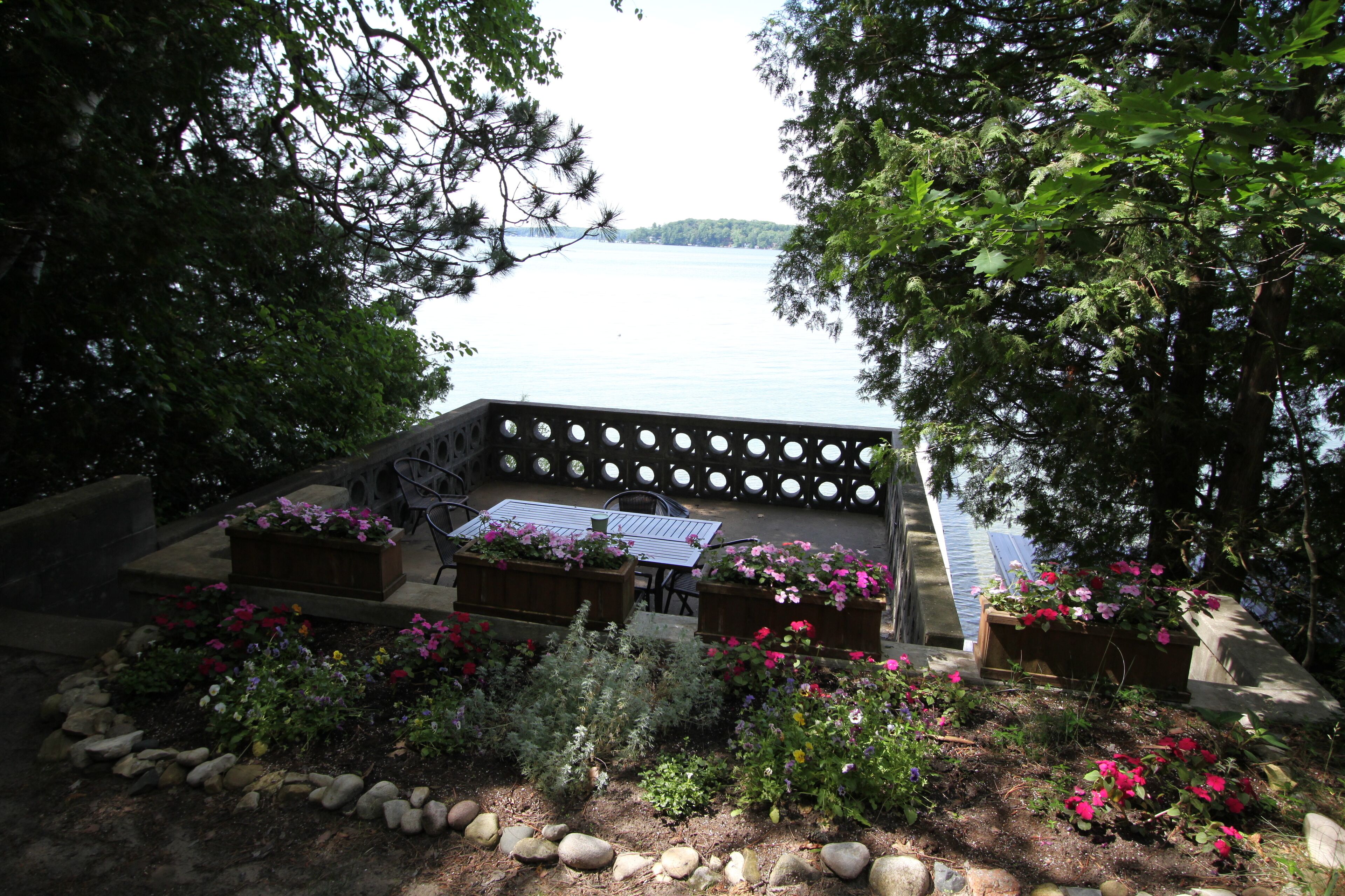 A patio with a table and chairs, surrounded by flower beds and a view of a lake.