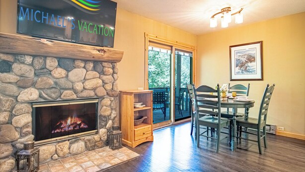 A cozy living room with a stone fireplace, a dining area with a table and chairs, and a large window offering a view of the outdoors. - SV113 - Walk to Lifts & Town - Hot Tub & Pool (Ketchum)