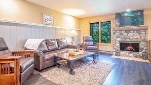 A cozy living room with a stone fireplace, leather sofas, and a large window offering a view of the outdoors. - SV113 - Walk to Lifts & Town - Hot Tub & Pool (Ketchum)