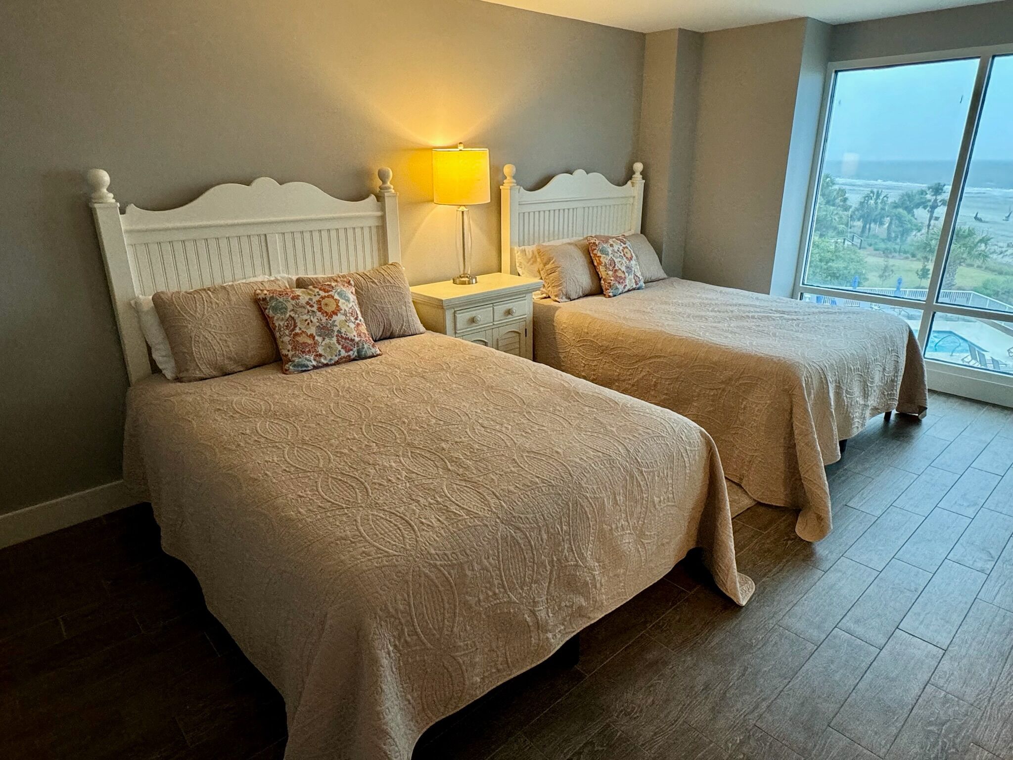 A hotel room with two beds, a nightstand, a lamp, and a large window with a view.