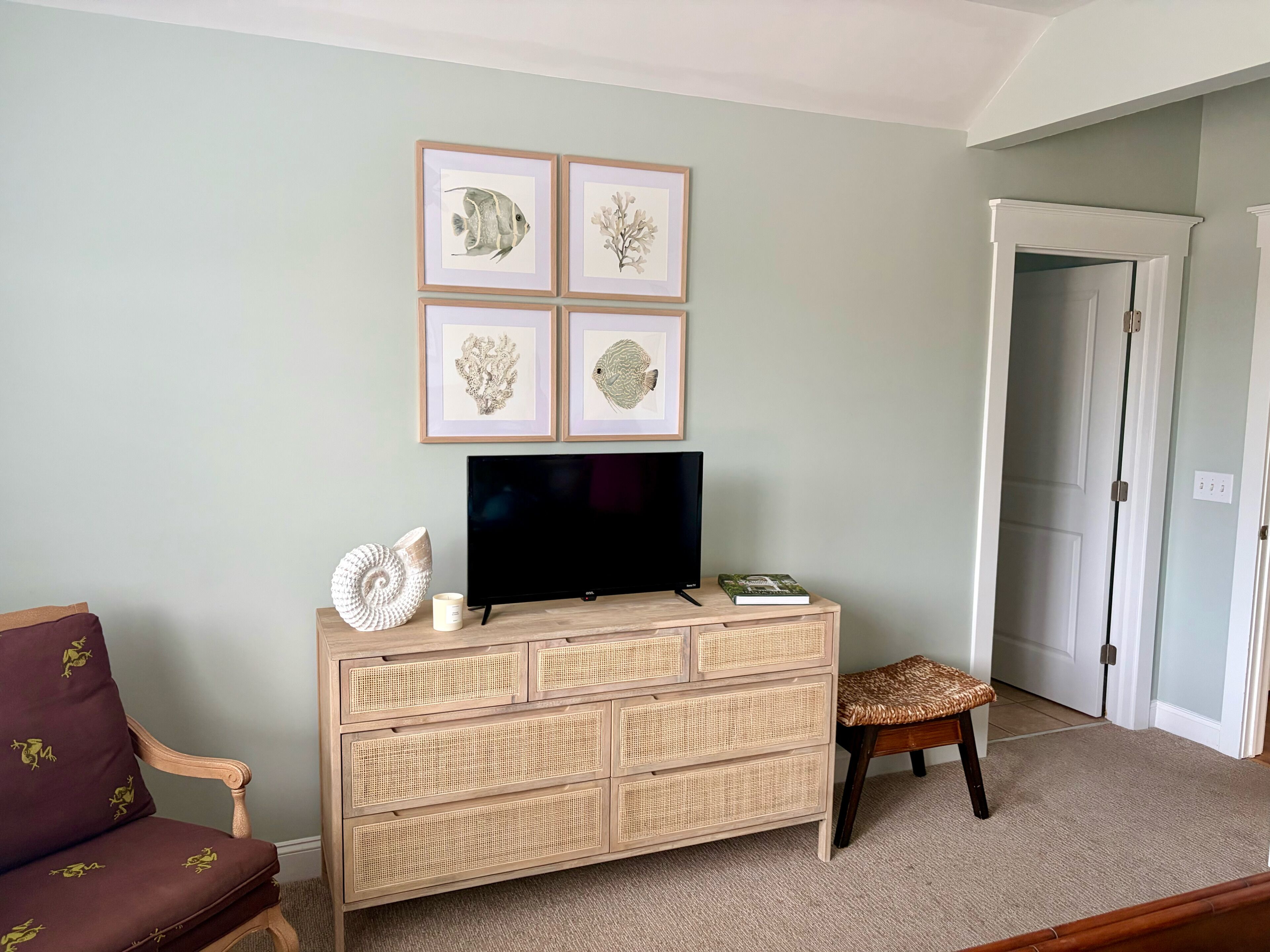 A living room with a television on a wooden dresser, framed artwork on the wall, a chair, and a door leading to another room.