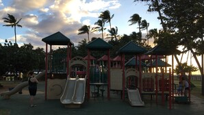 Un parque infantil con toboganes y estructuras para escalar, rodeado de palmeras y un cielo nublado.