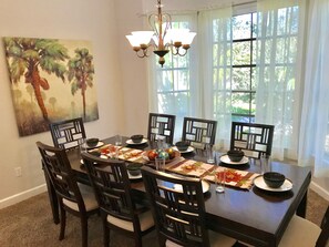 A dining room with a large table set for six, surrounded by dark wooden chairs. There is a chandelier above the table, and a window with sheer curtains allows natural light to enter. A painting of palm trees is visible on the wall. - Book now for spring break specials. Over 25 restaurants and bars open.  (Fort Myers Beach)