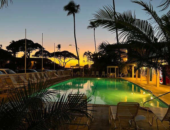 A pool area with lounge chairs and palm trees, surrounded by buildings with warm lighting. - Guests Love Our Condo. You Will Too! (Kihei)