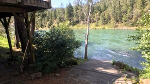 Une terrasse en bois donnant sur une rivière, entourée d’une forêt dense et sous un ciel dégagé.