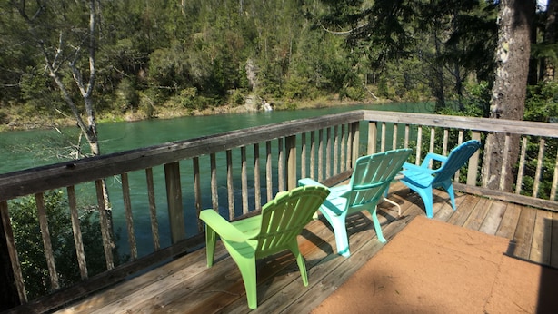 Une terrasse en bois avec deux chaises vertes donnant sur une rivière, entourée d’arbres et sous un ciel dégagé.