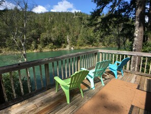 A wooden deck with two green chairs facing a river, surrounded by trees and a clear sky.