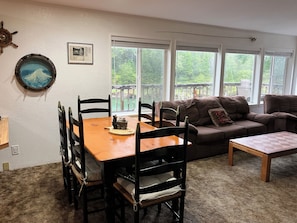 A dining area with a wooden table and chairs, a brown sofa, and a window with a view of greenery.