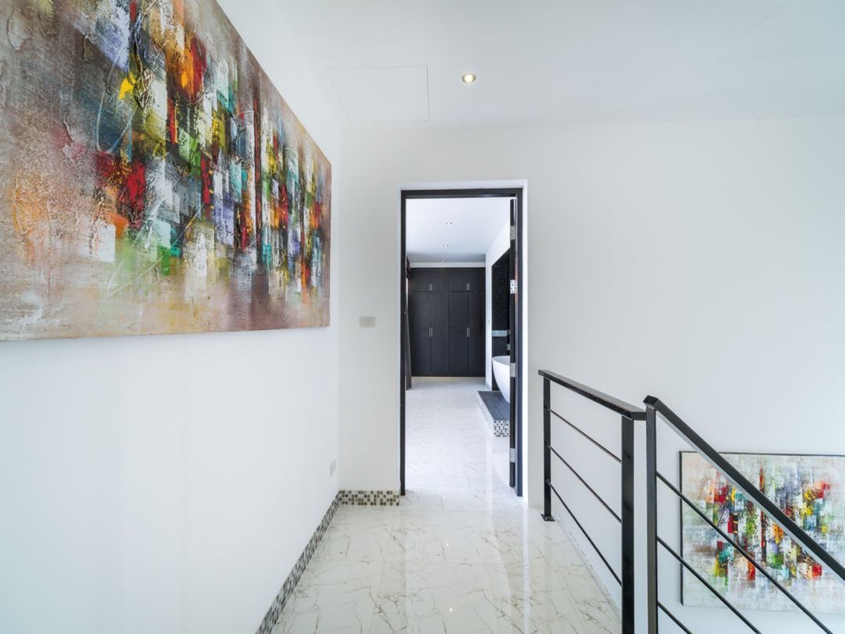 Un couloir avec une grande peinture abstraite sur le mur de gauche, un escalier avec une rampe en métal sur le côté droit, et une porte sombre au fond.