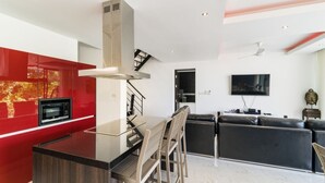 A modern kitchen with a central island, a dining area with bar stools, a black sofa, and a flat-screen TV mounted on the wall. - 3 Bedroom Sea View Villa 2 - Chaweng Noi SDV162-By Samui Dream Villas (Koh Samui)