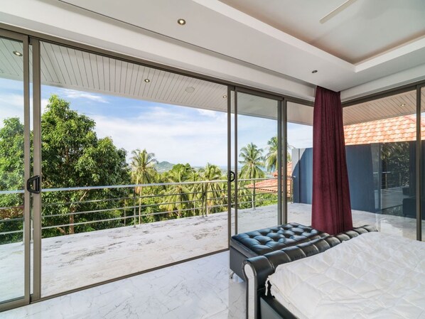 A modern bedroom with a large sliding glass door leading to a balcony with a view of palm trees and a distant mountain.