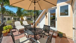 Une terrasse avec une table ronde, des chaises et un grand parasol. On y trouve également des plantes en pot et un escalier menant à un niveau supérieur.