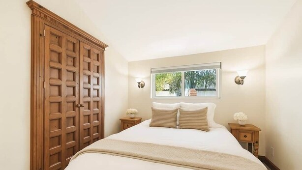 Une chambre avec une grande armoire en bois, un lit avec une literie blanche, deux tables de chevet avec des lampes et une fenêtre donnant sur un paysage verdoyant.