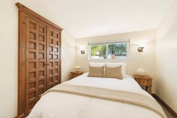 Ein Schlafzimmer mit großem Holzschrank, einem Bett mit weißer Bettwäsche, zwei Nachttischen mit Lampen und einem Fenster mit Blick ins Grüne.