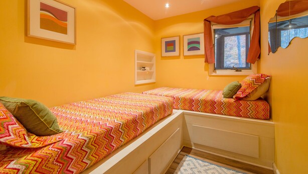 A bedroom with two beds, a window with curtains, a shelf, and artwork on the walls.