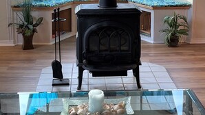 A cozy room with a black wood-burning stove, large windows with blue curtains, and a glass table with seashells. - Romantic Riverfront Retreat with Covered Deck & Fire Pit-walk to Beach! (Seaside)