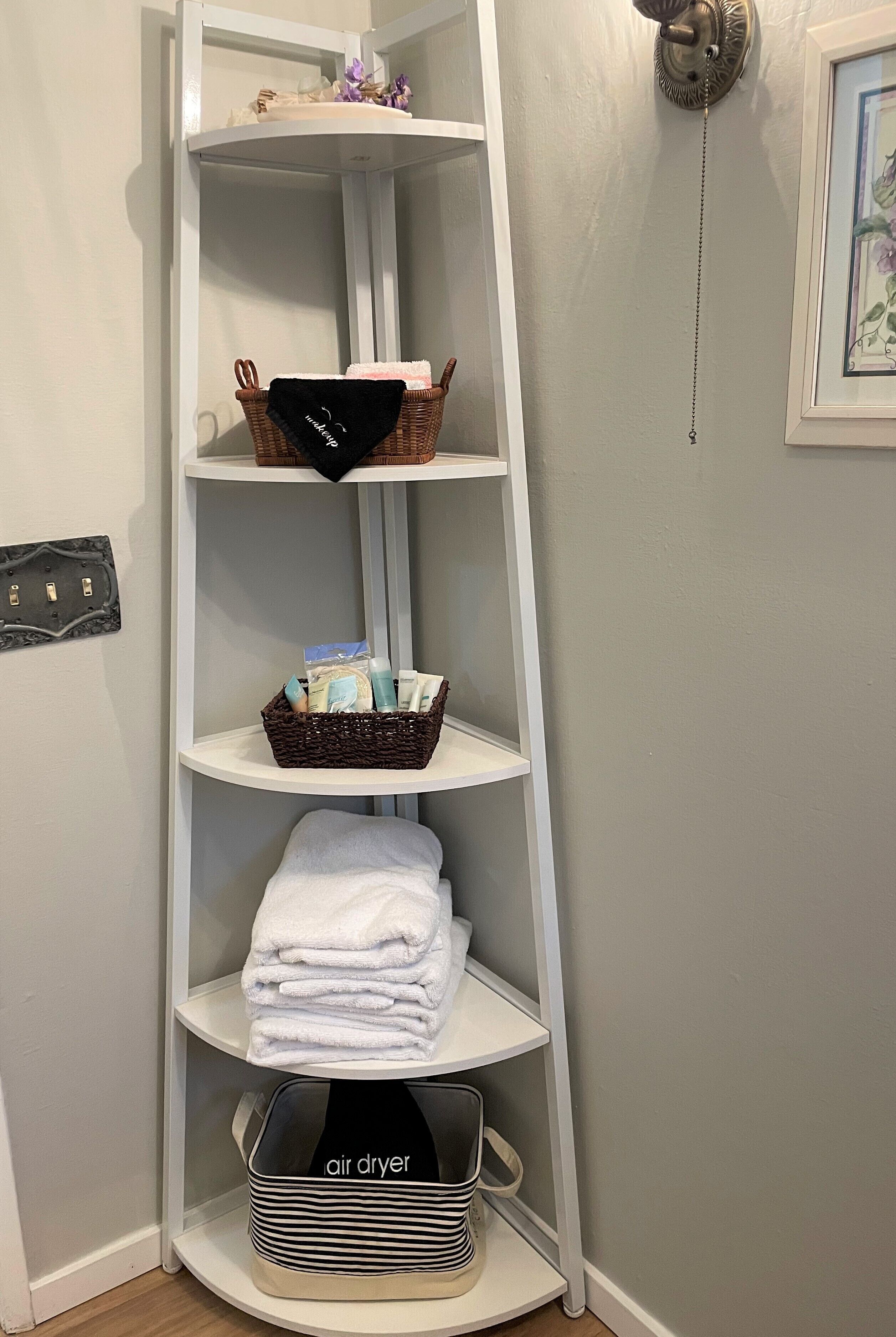 Un meuble d’angle pour le rangement, doté d’étagères où sont rangés des paniers, des serviettes et un sèche-cheveux.