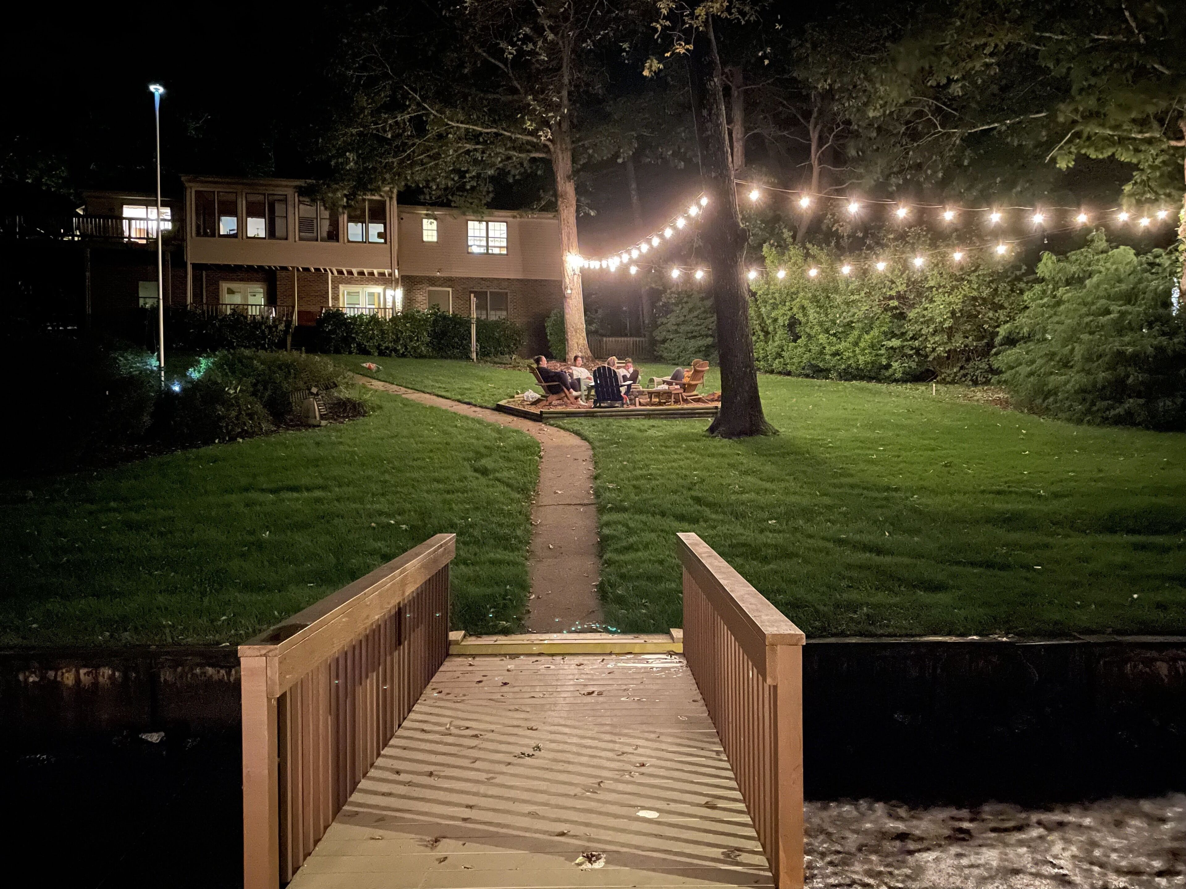 A wooden deck leading to a grassy area with string lights, a building in the background, and a tree.