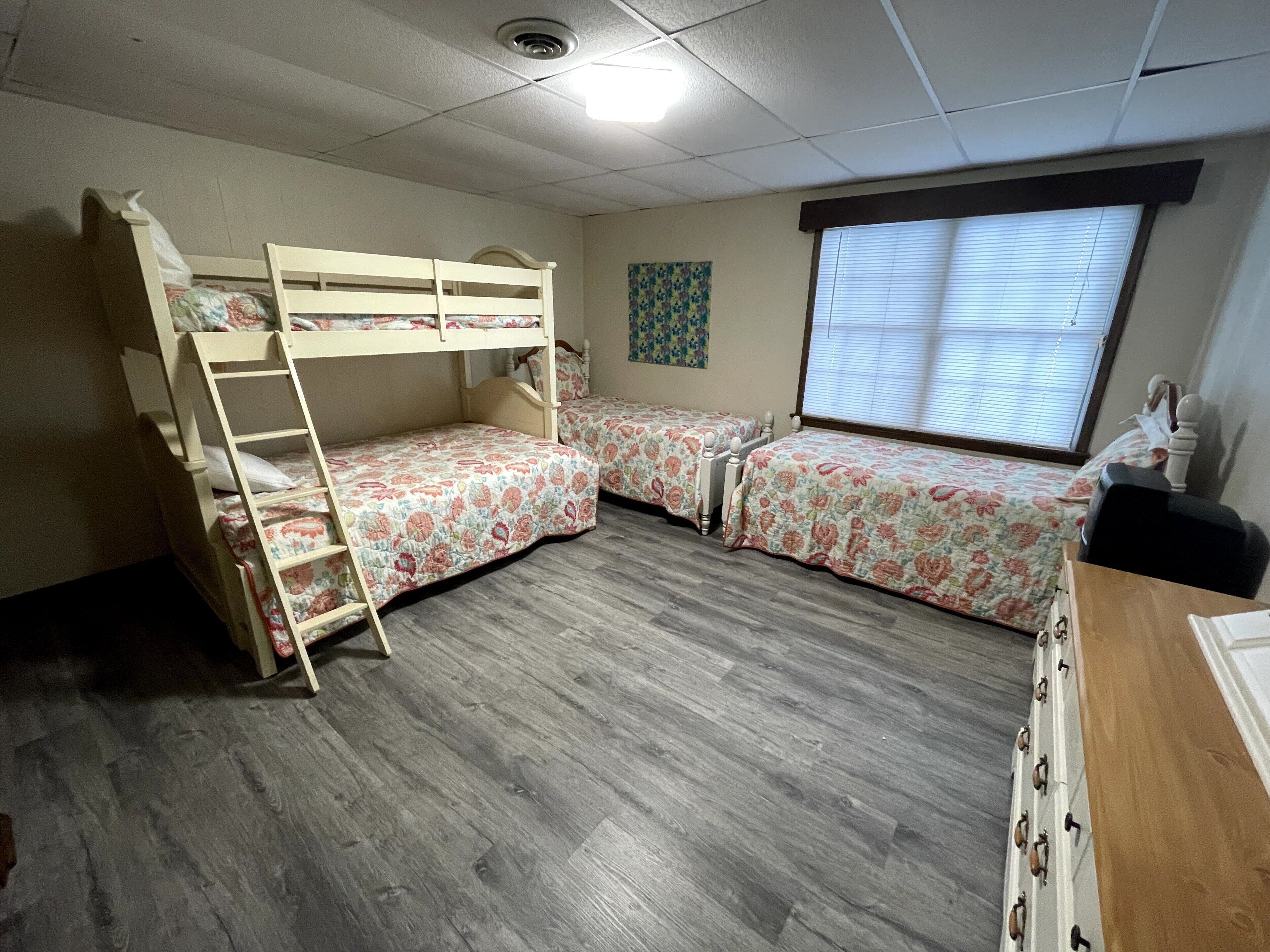 A room with bunk beds, a window with blinds, and a wooden dresser.