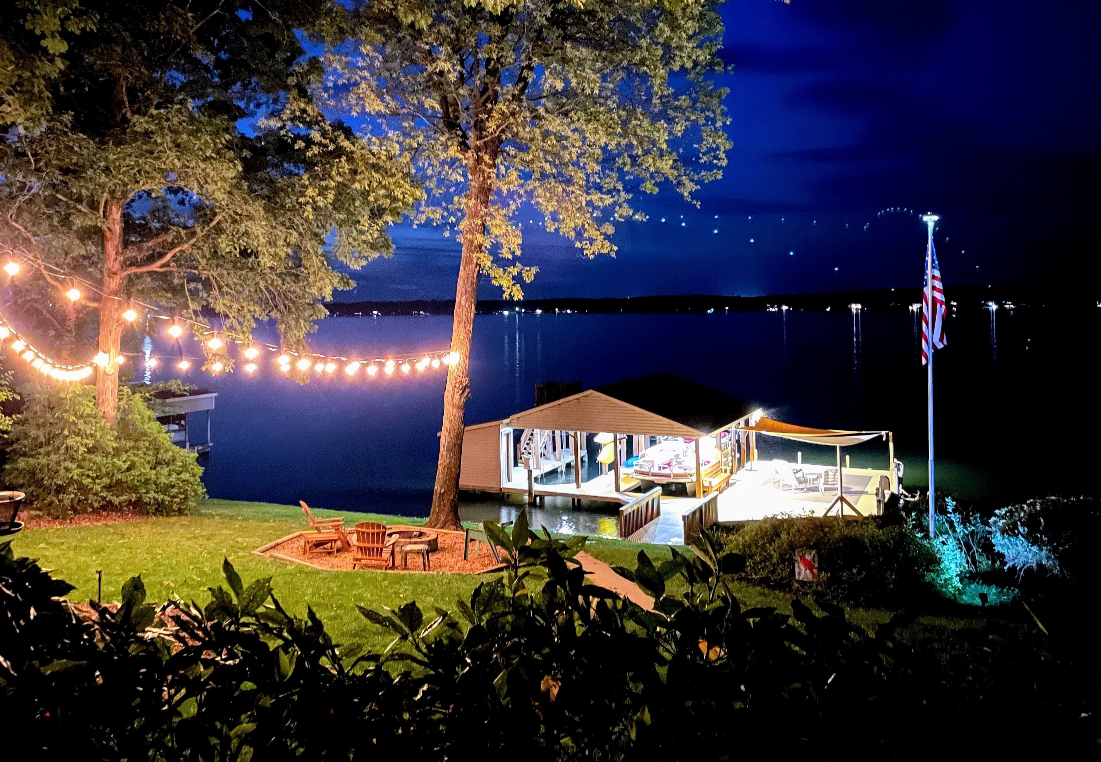 Un chalet au bord du lac, décoré de guirlandes lumineuses, avec un drapeau et une terrasse offrant une vue sur l’eau.