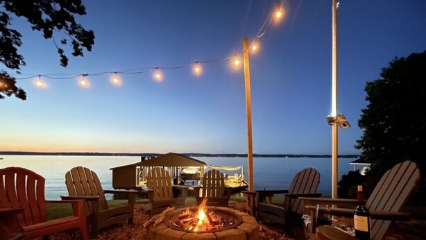Un foyer entouré de chaises Adirondack, une table et des guirlandes lumineuses, le tout sur fond d’un lac au crépuscule.