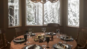 Ein Esszimmer mit einer Chandelüster, einem runden Tisch für vier Personen und großen Fenstern mit Blick auf einen verschneiten Wald.