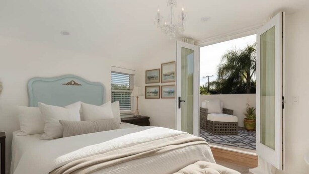 A bedroom with a bed, a nightstand, a chandelier, a window with blinds, and a door leading to an outdoor area with a chair and a plant.