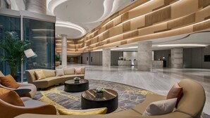 A spacious lobby with a modern design, featuring a central seating area with sofas and a coffee table, a reception desk, and a large plant.