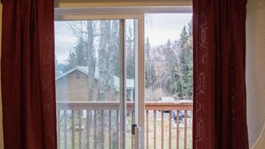 A room with a bed, a window with curtains, and a view of a deck and trees outside.