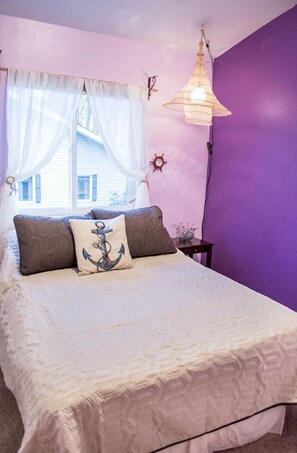 A bedroom with a bed, a window with curtains, a hanging light fixture, and a wall clock.