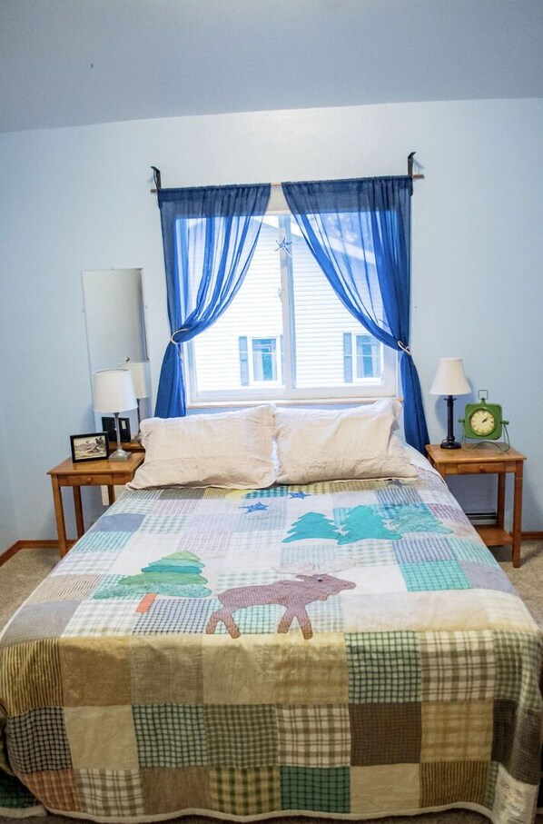 A bedroom with a bed, two nightstands, a window with blue curtains, and a green clock on one of the nightstands.