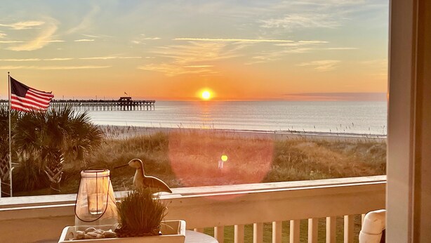Un balcon offrant une vue sur le coucher de soleil sur l’océan, une jetée visible au loin, et une table agrémentée d’une bougie et d’objets décoratifs.