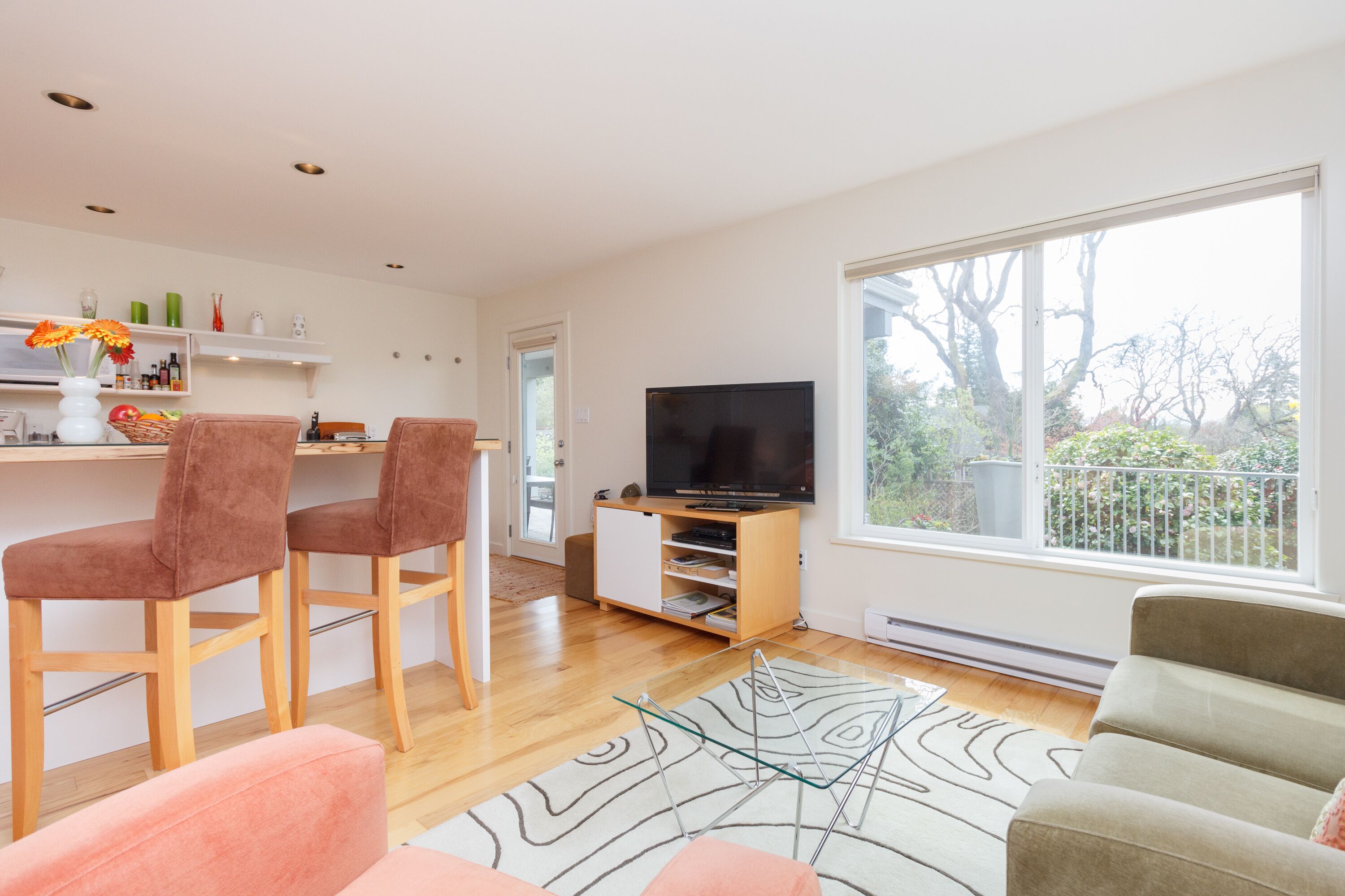 A modern living room with a large window, a flat-screen TV, and a kitchen area with bar stools.