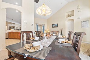 A dining room with a large table set for six, surrounded by wooden chairs, a chandelier, and a kitchen in the background.