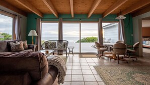 A spacious living room with a large sofa, a coffee table, and a dining area with chairs. The room has a wooden ceiling and large windows offering a view of the ocean.