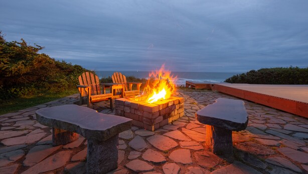 A stone fire pit with chairs and a bench, overlooking the ocean.