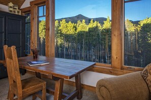 Una mesa de comedor de madera con sillas, una pequeña planta en maceta y un libro. Amplias ventanas ofrecen vistas a un bosque y una montaña.