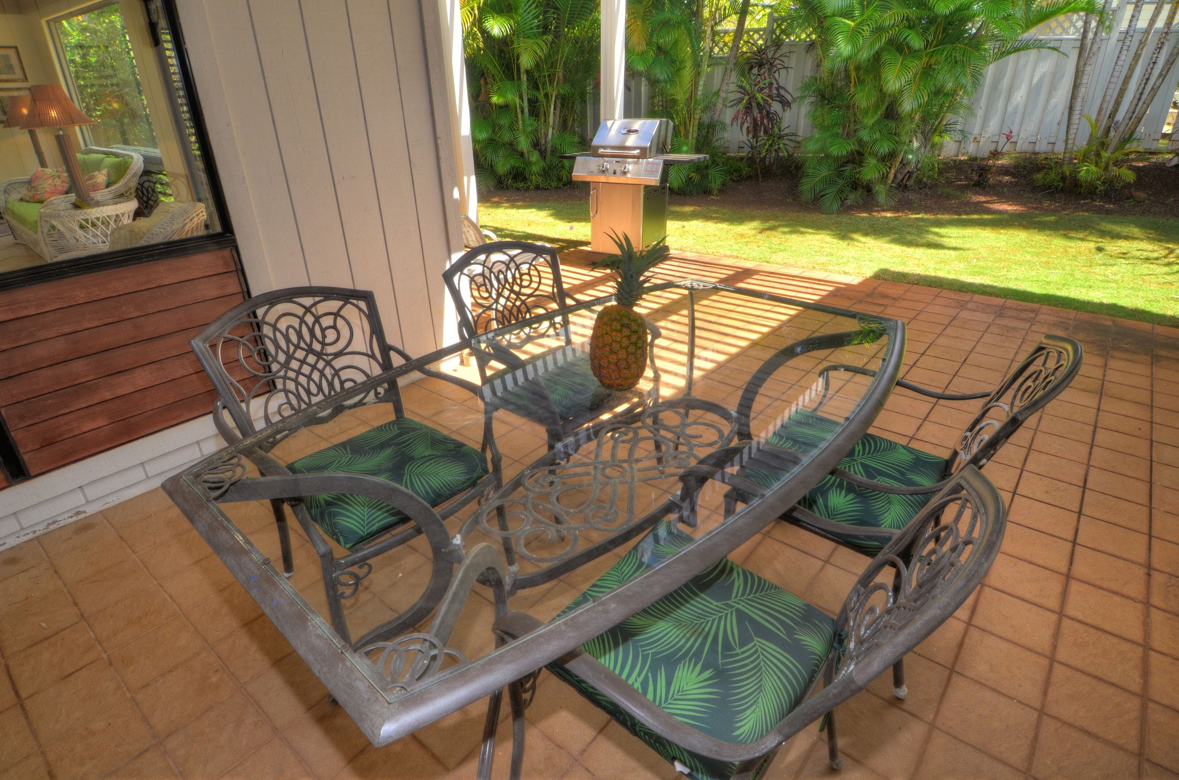 Eine Terrasse mit einem Metalltisch und Stühlen, einer Ananas auf dem Tisch und einem Grill im Hintergrund.