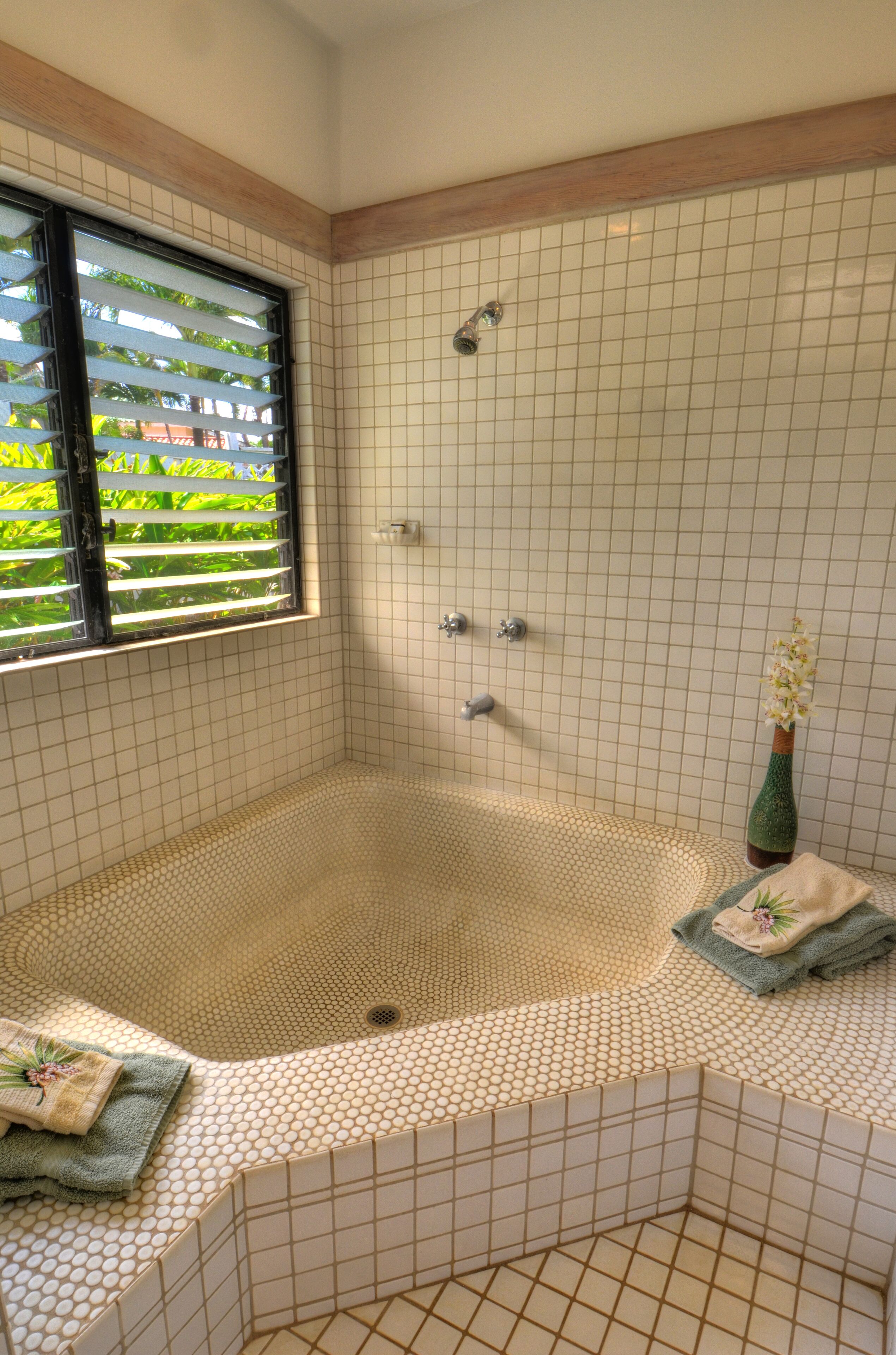 A bathroom with a large bathtub, a window with blinds, and a small vase with flowers.