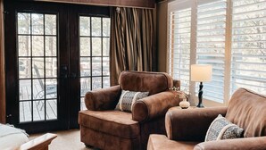 A cozy living room with a brown leather sofa, a wooden coffee table, and a lamp. There is a large window with blinds and a view of trees outside. - Timberloft Luxury Lodge, HOT TUB , GOLF COURSE views, secluded & private (Flagstaff)