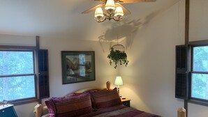 A bedroom with a bed, a ceiling fan, a window with shutters, a framed picture, and a hanging plant.