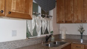 Eine Küche mit Granit-Arbeitsplatten, Holzschränken, einer Edelstahl-Spüle und einem Fenster mit weißen Vorhängen.