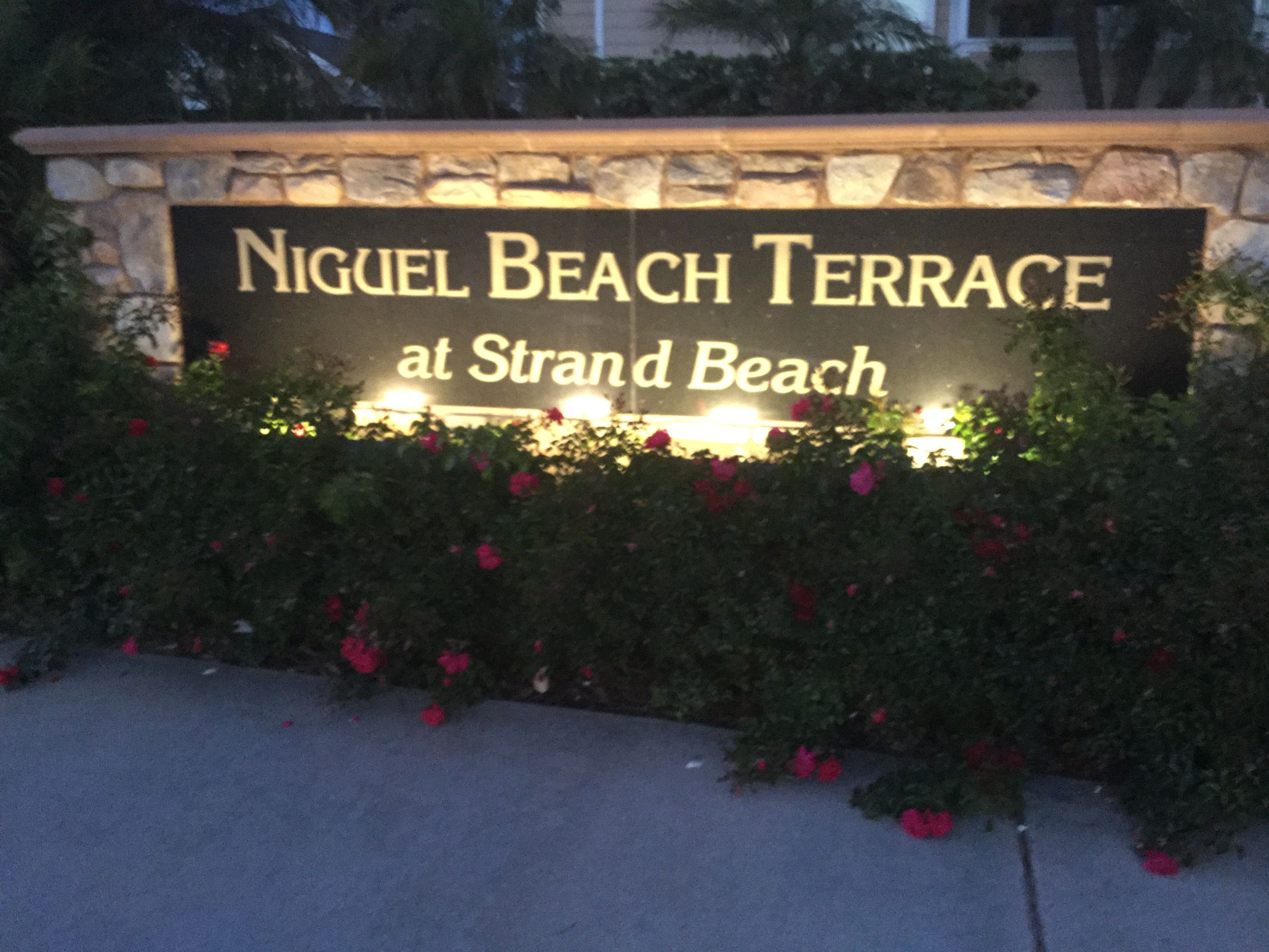 A sign for 'Nigel Beach Terrace at Strand Beach' with a stone wall and flowering plants.