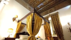 A four-poster bed with yellow curtains, a wooden headboard, and a white bedspread. There is a nightstand with a lamp, a chair, and a mirror on the wall. - Residenza Sinibaldi - XVI century experience (Rome)