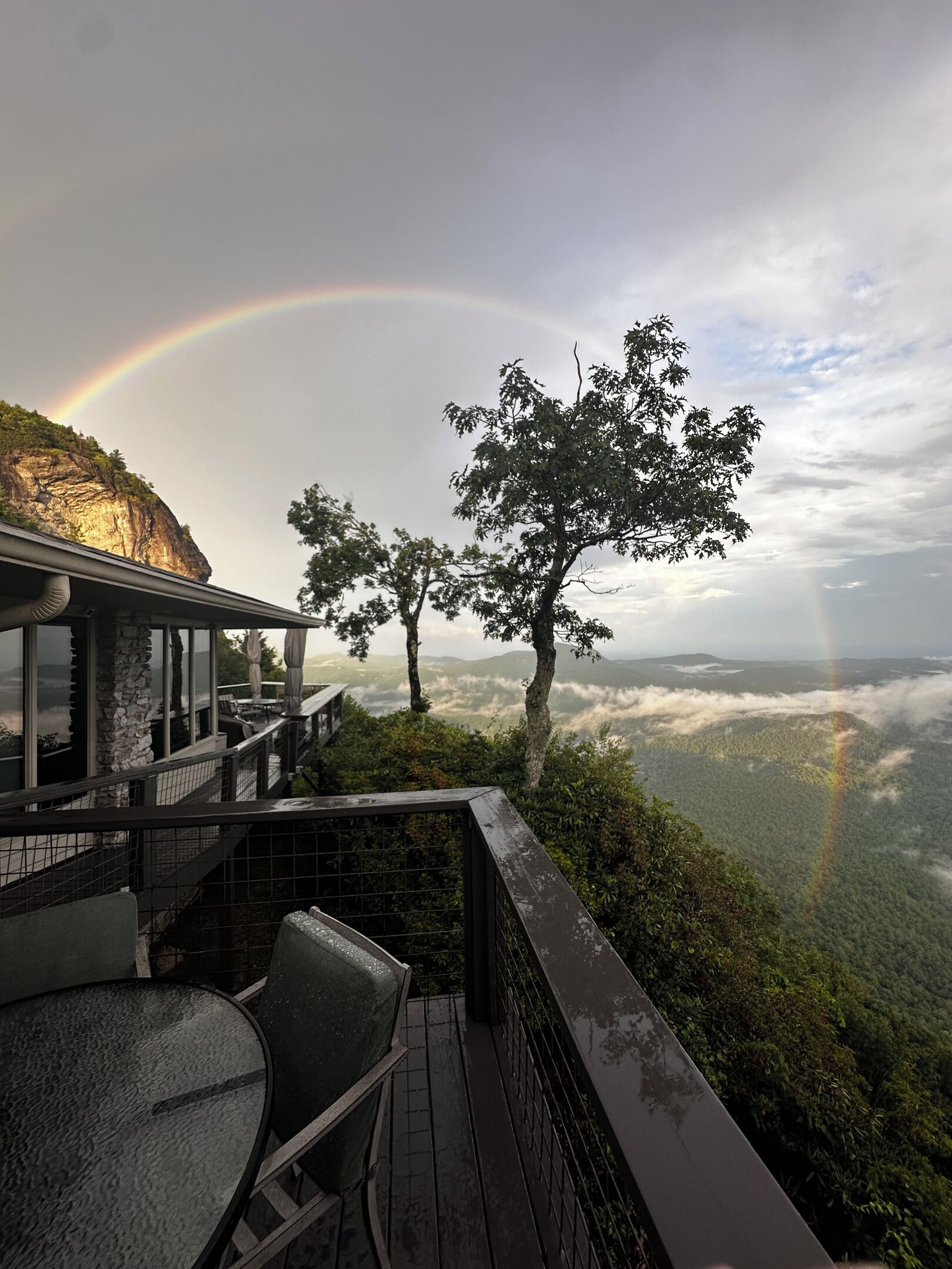 A wooden deck with a glass balustrade overlooking a mountainous landscape with a rainbow.