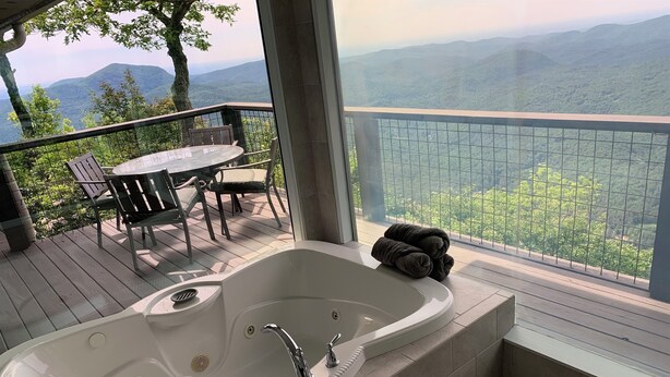 En balkon med et hvidt badekar, et bord med stole og udsigt til et bjerglandskab.