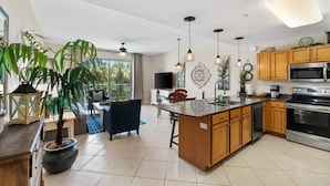 A modern kitchen with wooden cabinets, a large island, stainless steel appliances, and a dining area with a blue sofa and a TV. - Beach Resort 215 Beach Setup included* HUGE value added.  Perfect Pool View. Convenient Access (Miramar Beach)