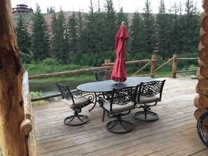 Una terraza de madera con una mesa redonda y cuatro sillas, un paraguas y vistas a un río y colinas boscosas.