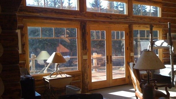 A log cabin interior with large windows, a wooden table, and a lamp. - WindRiver/ Yellowstone Cabin (Dubois)