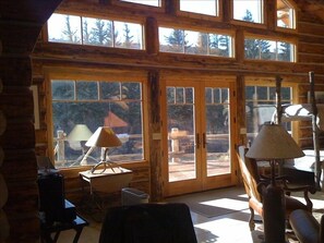 Interior de una cabaña de troncos con ventanas amplias, una mesa de madera y una lámpara.